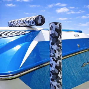 May include: Two boat accessories with a camouflage pattern in shades of gray and black. One is cylindrical and horizontal, the other is vertical. The accessories are on a blue and white boat, with a blue sky in the background.