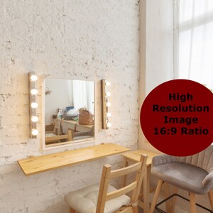 May include: A white vanity mirror with light bulbs on either side, a wooden countertop, and two wooden chairs. A plant is in a pot on a windowsill behind the vanity.