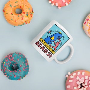 K&ouml;nnte beinhalten: Wei&szlig;e Keramiktasse mit einer blauen und gelben Grafik einer Person, die ein Polizeiauto umdreht. Der Text "ACAB20" ist auf die Tasse gedruckt.