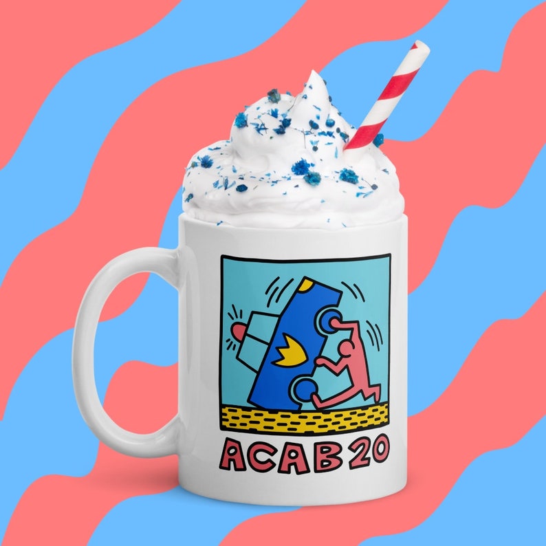 K&ouml;nnte beinhalten: Eine wei&szlig;e Keramiktasse mit einer blauen und roten Grafik einer Person, die ein Polizeiauto umkippt. Die Grafik ist von einem schwarzen und gelben Rand umgeben. Der Text "ACAB20" ist in Schwarz unter der Grafik gedruckt. Die Tasse ist mit Schlagsahne und blauen Streuseln bedeckt und hat einen rot-wei&szlig; gestreiften Strohhalm.
