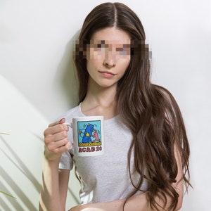 K&ouml;nnte beinhalten: Eine wei&szlig;e Keramiktasse mit einer blauen und roten Illustration einer Person, die ein Auto schiebt. Der Text "ACAB20" ist auf der Unterseite der Tasse aufgedruckt.