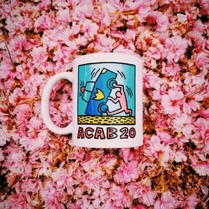 K&ouml;nnte beinhalten: Wei&szlig;e Keramiktasse mit einer blauen, gelben und roten Grafik einer Person, die &uuml;ber ein Auto springt. Der Text "ACAB 20" ist auf die Tasse gedruckt.