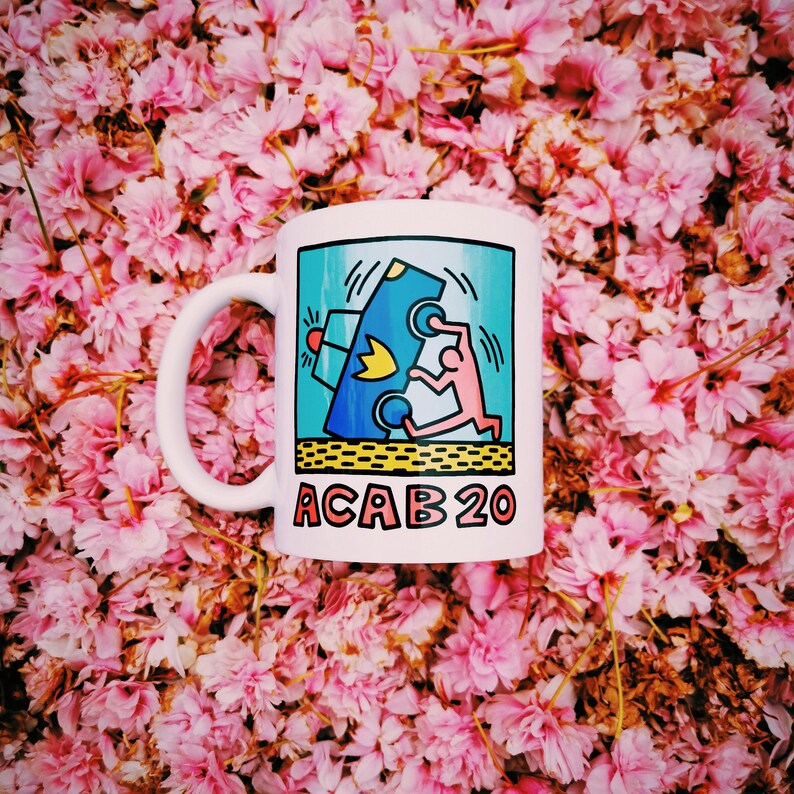 K&ouml;nnte beinhalten: Wei&szlig;e Keramiktasse mit einer blauen, gelben und roten Grafik einer Person, die &uuml;ber ein Auto springt. Der Text "ACAB 20" ist auf die Tasse gedruckt.