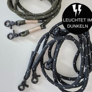 Op de afbeelding: Twee hondenriemen, een zwarte en een groene, met reflecterende accenten. Beide riemen hebben zwarte metalen clips en de zwarte riem heeft de tekst "LEUCHTET IM DUNKELN" op een zwarte cirkel met een bliksem.