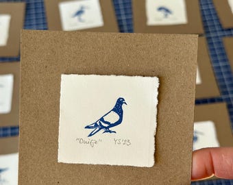 Op de afbeelding: Een handgeprinte illustratie van een blauwe en witte duif op een witte kaart met de tekst "Duifje" en "YS'23" in blauwe inkt. De kaart is gemonteerd op een bruine kaart.