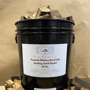 May include: A black bucket filled with 20 pounds of whiskey barrel oak smoking chunks. The bucket has a white label with the text "Koch Barrel Co. Premium Whiskey Barrel Oak Smoking Chunk Bucket 20 lbs. www.kochbarrelco.com".