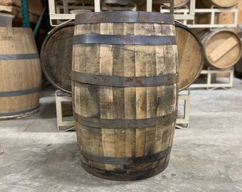 May include: A rustic wooden barrel with dark metal hoops. The barrel is constructed from vertical wooden staves and has a gently curved form. Other barrels are visible in the background, suggesting a distillery or storage environment.