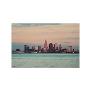 May include: A scenic view of a city skyline with various buildings, including skyscrapers and structures with spires, set against a backdrop of a serene body of water and a cloudy sky. The buildings are in shades of brown and red.
