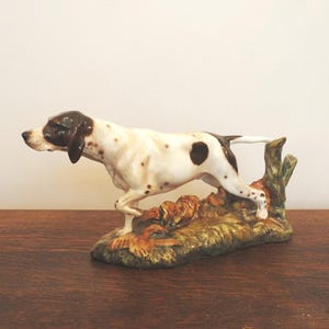 Puede incluir: Figura de cerámica de un perro Pointer en pose de caza. El perro es blanco con manchas marrones y cabeza marrón. Se encuentra sobre una base de follaje verde y marrón con un tronco de árbol. La figura está sobre una superficie de madera.