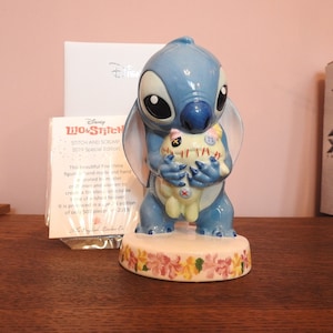 May include: A blue and white ceramic figurine of Stitch from the Disney movie Lilo & Stitch. Stitch is holding a small, white plush toy with red stitching. The figurine is sitting on a base with a floral design. The text on the card reads "Disney Lilo & Stitch Stitch and Scrump 2019 Special Edition".