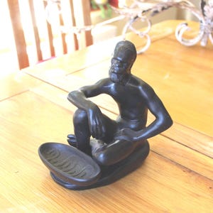 May include: A matte black figurine of a seated person with a beard, leaning forward with one arm resting on a knee. The figure is attached to a small, oval-shaped tray with a textured, wavy pattern on the interior surface, crafted as a single piece.