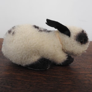 Puede incluir: Un conejito de peluche con cuerpo color crema y manchas negras. El conejo tiene orejas y patas negras. Está sobre una superficie de madera.