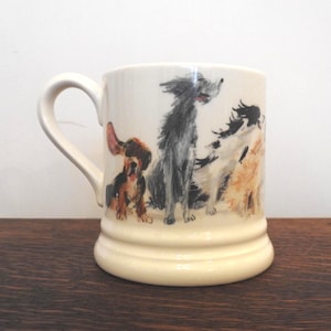 May include: Cream-coloured ceramic mug with a hand-painted illustration of various dog breeds. The dogs are in black, brown, and tan. The mug has a curved handle and a slightly flared base. The mug is suitable for tea or coffee.
