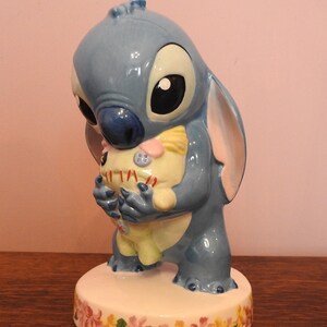 May include: A blue ceramic figurine of Stitch from Lilo & Stitch holding a plush toy. The figurine is standing on a white base with a floral design.