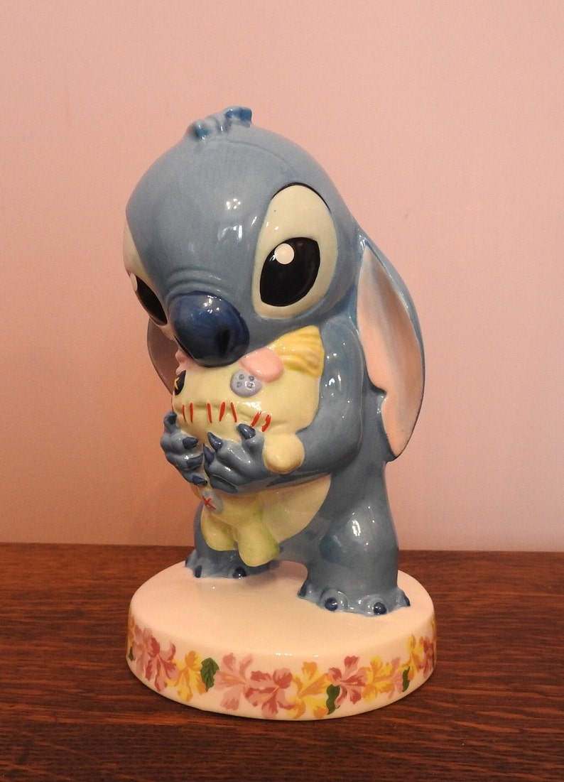 May include: A blue ceramic figurine of Stitch from Lilo & Stitch holding a plush toy. The figurine is standing on a white base with a floral design.
