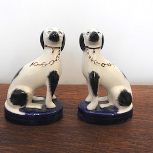 Op de afbeelding: Twee keramische hondenbeeldjes. De honden zijn wit met zwarte vlekken en zwarte oren. Ze hebben goudkleurige kettinghalsbanden en zitten op blauwe sokkels. De beeldjes staan op een houten oppervlak.