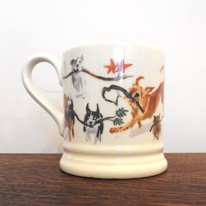 May include: Cream-colored ceramic mug featuring a playful dog illustration. The design includes various dog breeds, each holding a stick or branch. The mug has a sturdy handle and a slightly flared base, perfect for enjoying your favorite beverage.