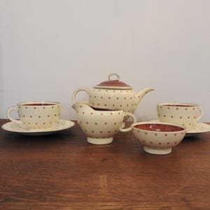 May include: A cream-coloured tea set with red polka dots. The set includes a teapot, creamer, sugar bowl, and two teacups with saucers. The teapot lid and the interior of the cups and bowl are a darker red. A classic tea set.