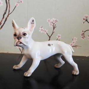 May include: A white porcelain figurine of a French Bulldog with black spots. The figurine is standing on a dark surface.