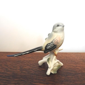 May include: A ceramic bird ornament perched on a white branch with green leaves. The bird features a grey head, a white body with pink hues, and a long black and white tail. The ornament sits on a wooden surface.