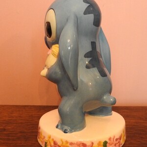 May include: A blue ceramic figurine of Stitch from Lilo & Stitch holding a small yellow and pink doll. The figurine is standing on a white base with a floral design.