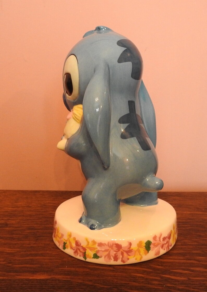 May include: A blue ceramic figurine of Stitch from Lilo & Stitch holding a small yellow and pink doll. The figurine is standing on a white base with a floral design.