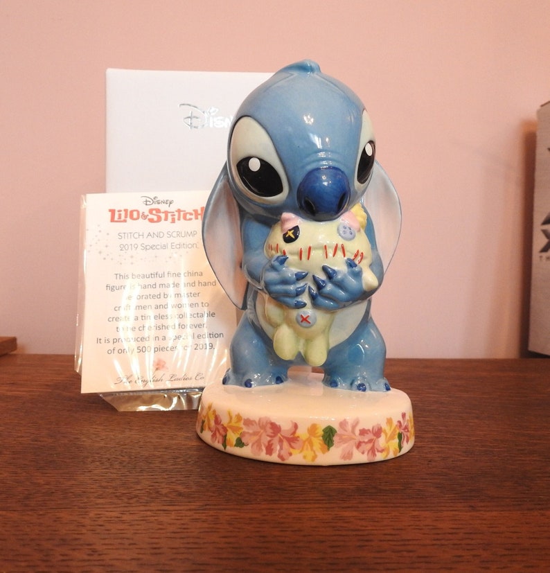 May include: A blue and white ceramic figurine of Stitch from the Disney movie Lilo & Stitch. Stitch is holding a small, white plush toy with red stitching. The figurine is sitting on a base with a floral design. The text on the card reads "Disney Lilo & Stitch Stitch and Scrump 2019 Special Edition".