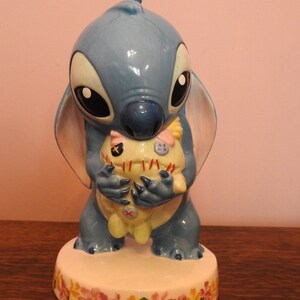 May include: A blue ceramic figurine of Stitch from Lilo & Stitch holding a yellow and white plush toy. The figurine is standing on a white base with a floral design.