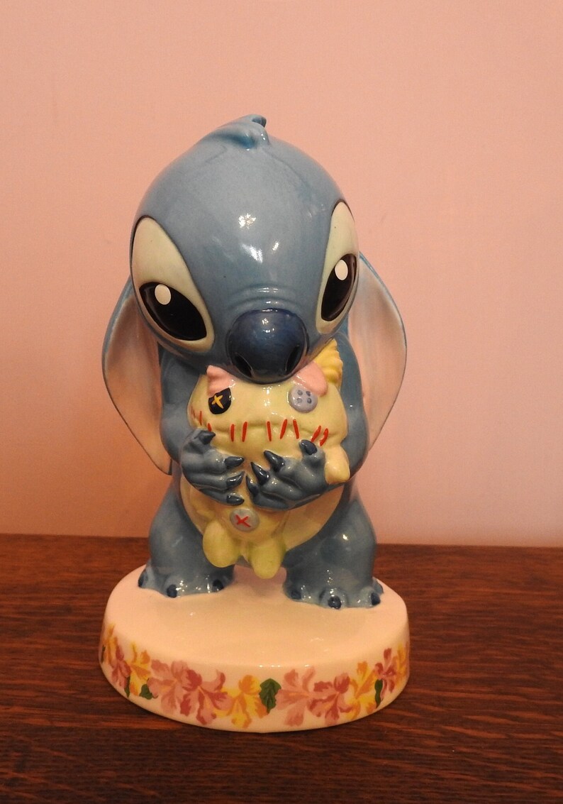 May include: A blue ceramic figurine of Stitch from Lilo & Stitch holding a yellow and white plush toy. The figurine is standing on a white base with a floral design.