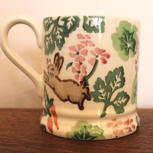 May include: A cream-coloured ceramic mug featuring a painted pattern of a brown rabbit, orange carrots, pink flowers, and green leaves. The mug has a curved handle on the left side and a slightly flared base, with a smooth, glossy finish throughout.