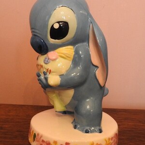 May include: A blue ceramic figurine of Stitch from Lilo & Stitch holding a small, yellow, and pink plush toy. The figurine is standing on a white base with a floral design.