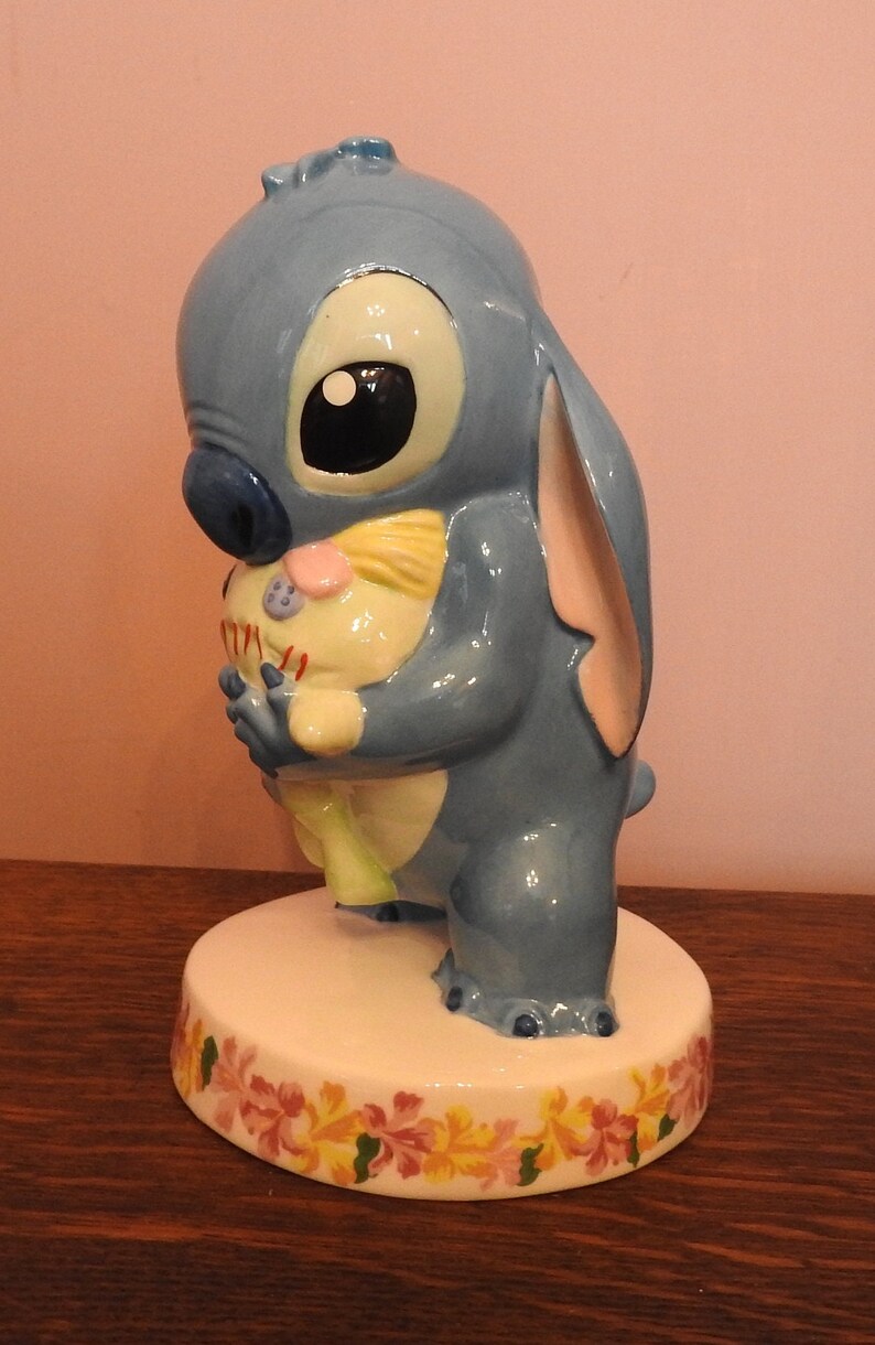 May include: A blue ceramic figurine of Stitch from Lilo & Stitch holding a small, yellow, and pink plush toy. The figurine is standing on a white base with a floral design.