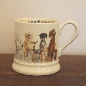 May include: A cream-colored ceramic mug featuring a wrap-around illustration of various dog breeds in shades of brown, white, black, and tan. The interior rim of the mug is decorated with a repeating pattern of small, dark-colored paw prints.