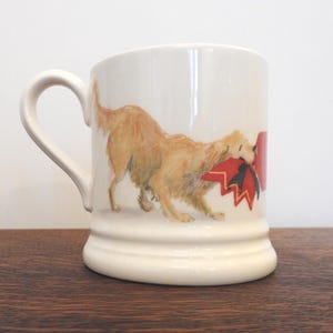 May include: Cream-colored ceramic mug featuring a golden retriever with a red bow in its mouth. The dog is depicted in a watercolor style, adding a touch of artistry to the design. The mug has a curved handle and a sturdy base.