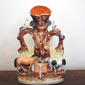 May include: A decorative ceramic vase featuring a tree-like structure with a large opening. Two birds perch on the branches, and a horse and cow stand at the base. The vase has a brown, orange, and cream color scheme.