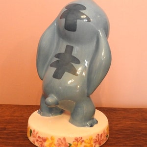 May include: A blue ceramic figurine of Stitch from Lilo & Stitch. The figurine is standing on a white base with a floral design. Stitch is in a playful pose with his arms raised and his head tilted back.