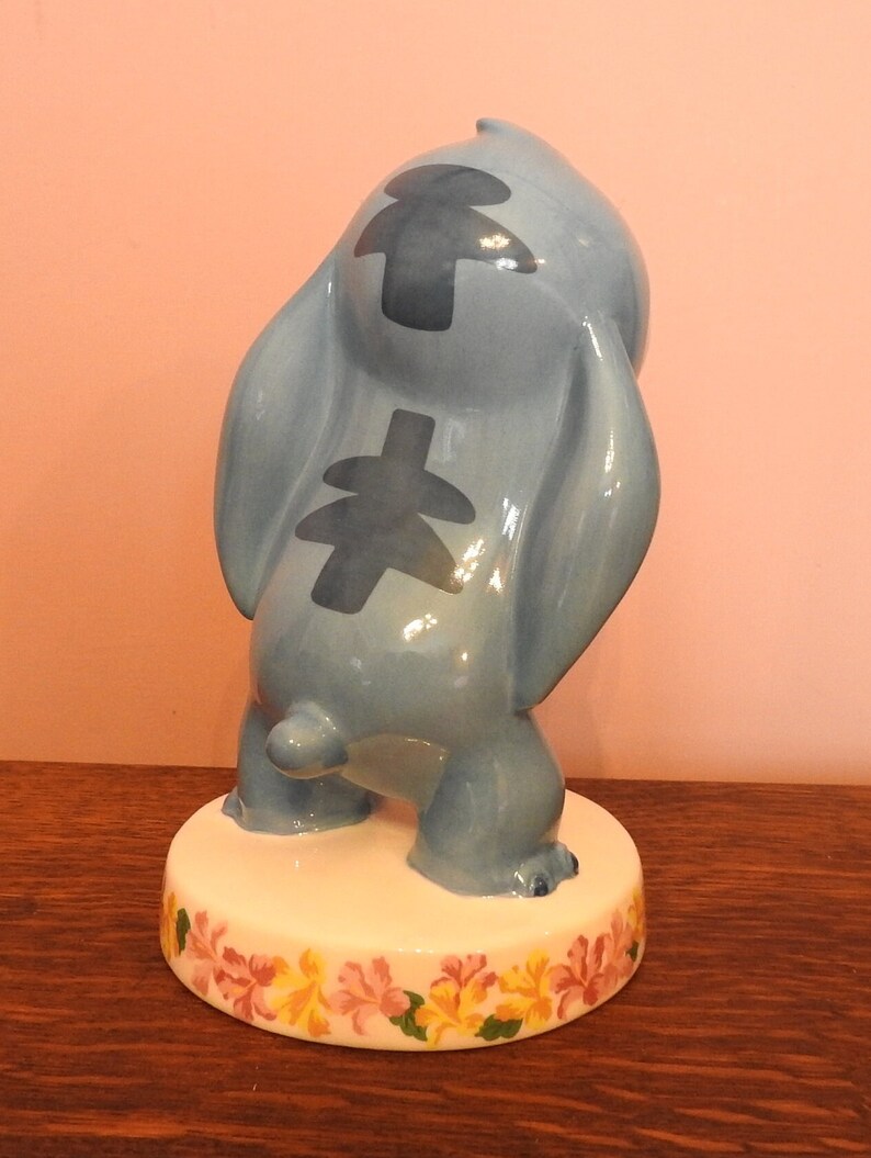 May include: A blue ceramic figurine of Stitch from Lilo & Stitch. The figurine is standing on a white base with a floral design. Stitch is in a playful pose with his arms raised and his head tilted back.