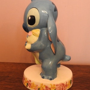 May include: A blue ceramic figurine of Stitch from Lilo & Stitch holding a yellow and pink plush toy. The figurine is standing on a white base with a floral design.