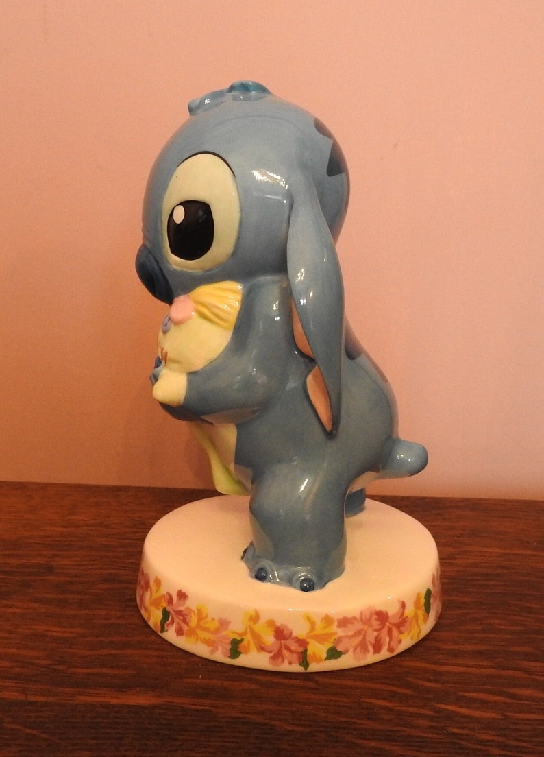 May include: A blue ceramic figurine of Stitch from Lilo & Stitch holding a yellow and pink plush toy. The figurine is standing on a white base with a floral design.
