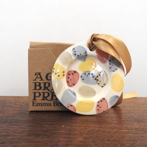 May include: A small, round, ceramic dish with a cream-colored base and colorful, abstract dot pattern in yellow, blue, and red. A gold ribbon is attached to the dish. The dish is next to a brown cardboard box with text.
