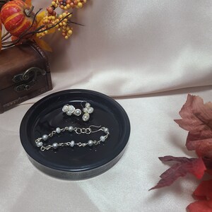 May include: A black ceramic dish with a pearl bracelet and pearl earrings. The dish has a decorative pattern around the edge.