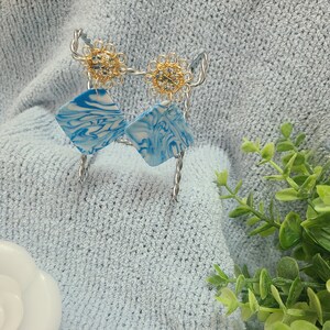 Puede incluir: Un par de pendientes de mármol azul y blanco con detalles dorados. Los pendientes están colgados de un soporte de alambre plateado.