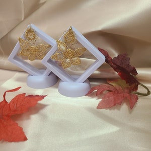 Puede incluir: Dos pares de pendientes de alambre dorado en forma de flor con un pequeño dije de flor que cuelga del centro. Los pendientes se muestran en dos cajas de terciopelo morado.