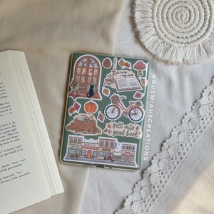 May include: A green e-reader case adorned with fall-themed stickers, including a bicycle, pumpkin, and the phrase "fall into a good book!". A book is open next to the e-reader, with a white lace trim and a macrame coaster in the background.