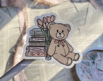Op de afbeelding: Een sticker met een bruine teddybeer die op een stapel boeken zit met een vaas met roze tulpen. De sticker heeft de tekst "@RUSMIAHSCREATIONS" onderaan.