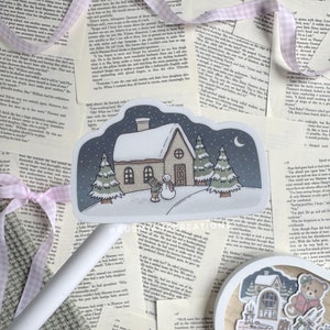 May include: A sticker featuring a winter scene with a small house, two pine trees, a snowman, and a person building the snowman. The sky is dark blue with a crescent moon and stars. The house has a white roof and a brown door. The sticker is on a white background.