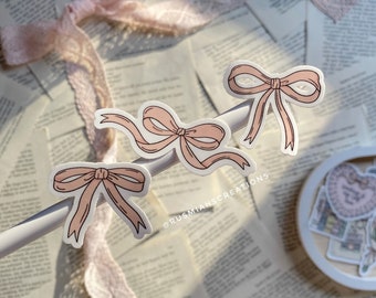 May include: Three pink bow stickers with black outlines are displayed on a white surface, set against a backdrop of vintage book pages and a pink lace ribbon. The stickers have a delicate, hand-drawn aesthetic.