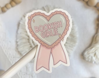 May include: A sticker with a heart-shaped badge that says "BOOKISH GIRLIE" in pink and white text. The badge has a pink ribbon and a light green background. The sticker is on a white surface.