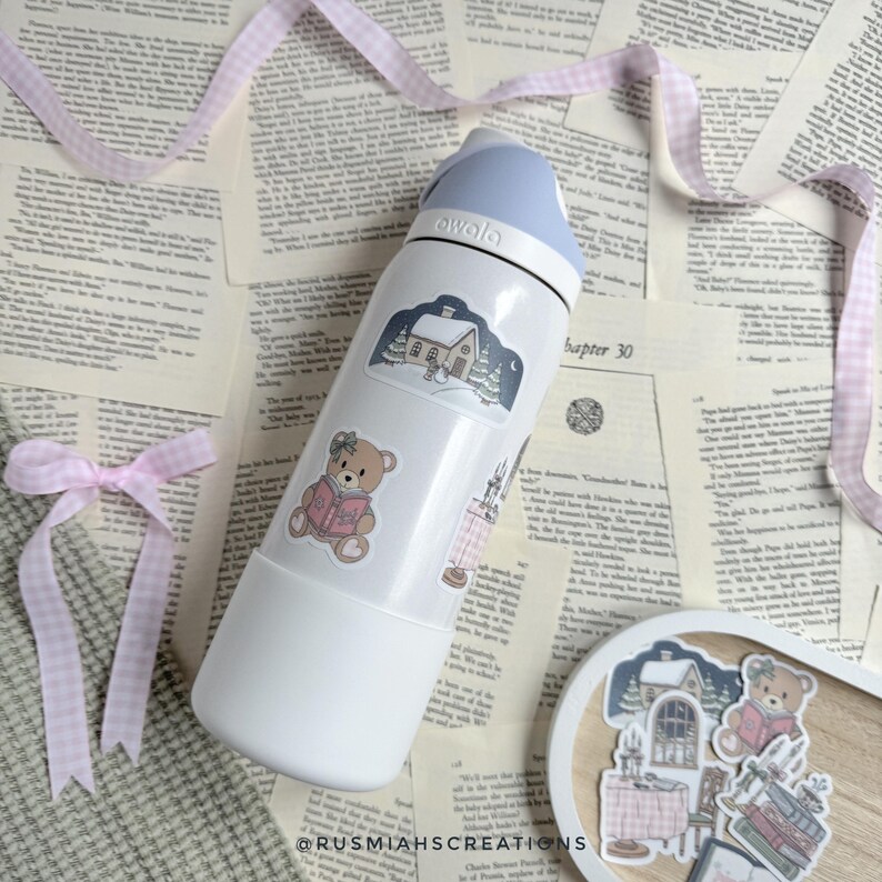 May include: White reusable water bottle with a blue lid and various stickers. The stickers feature a bear reading a book, a house with a snowy roof, and a winter scene. The bottle is on a surface with a pink and white checkered pattern.