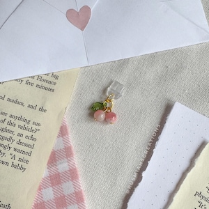 May include: A close-up of a pink and green cherry charm with a gold chain and a clear plastic clip. The charm is on a white and pink checkered background.